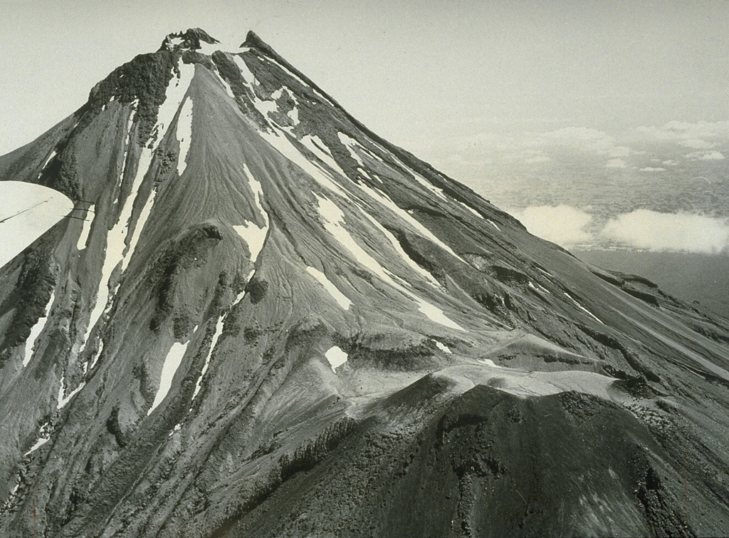 An aerial view from the south shows Taranaki, with its parasitic cone Fanthams Peak at the bottom right. The summit contains a partially destroyed lava dome and four other lava domes are located low on the S and N flanks. Taranaki collapsed and rebuilt itself repetitively during the late Pleistocene and Holocene. Photo by Jim Cole, 1971 (University of Canterbury).