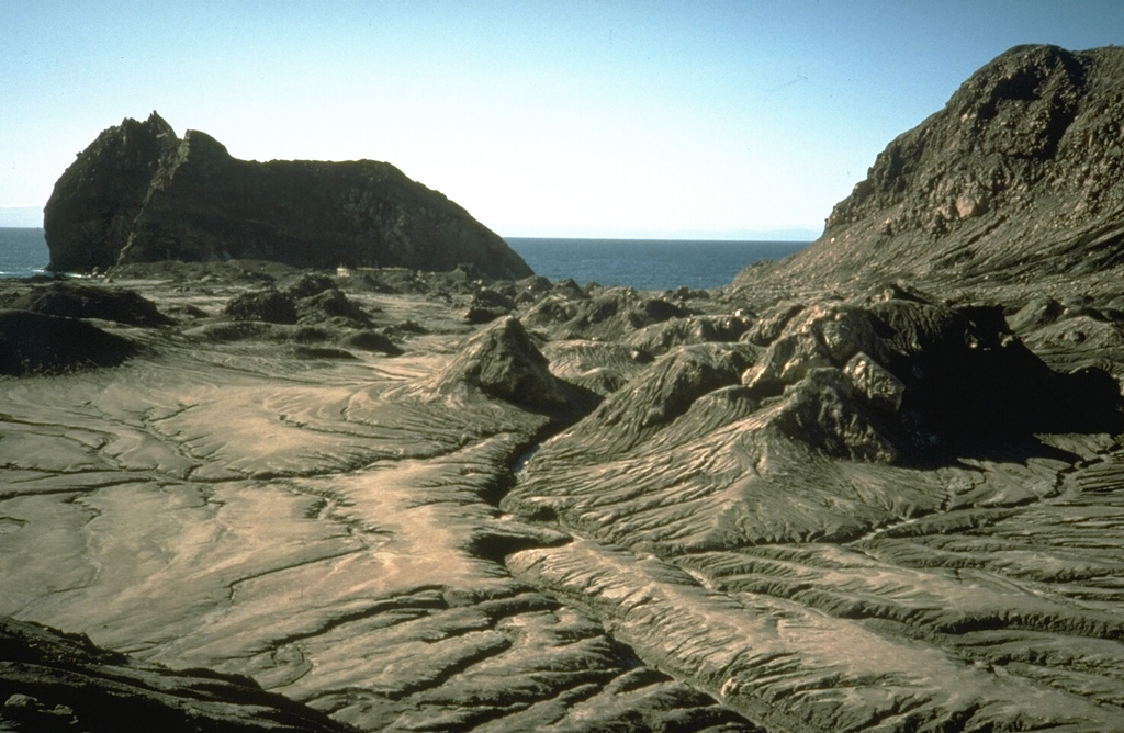 The small hills in the foreground are debris avalanche hummocks from a landslide on White Island in September 1914. The landslide occurred when part of the crater rim collapsed in the absence of any eruptive activity. The high-velocity avalanche swept across the crater floor, destroying the buildings of a sulfur-mining plant and burying 11 workers. The large hill in the background to the SE is not an avalanche hummock but a remnant of the outer crater wall. Photo by Richard Waitt, 1986 (U.S. Geological Survey).