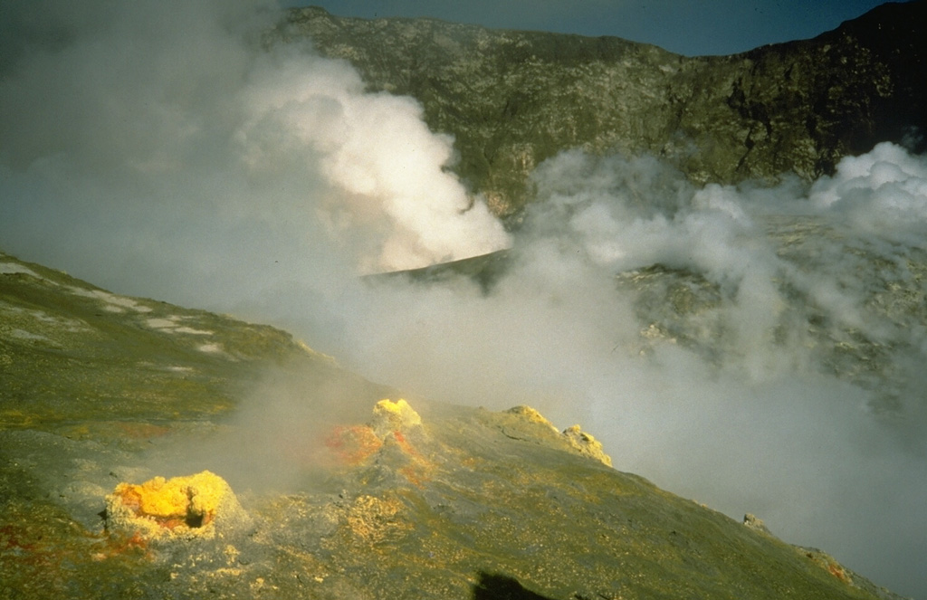 Sulfur deposits on the margins of fumarole vents on White Island volcano (also called Whakaari), with steam rising above a larger vent in the background. This photo was taken shortly after the beginning of an explosive eruption in February 1986 that lasted until July 1994. Photo by Richard Waitt, 1986 (U.S. Geological Survey).