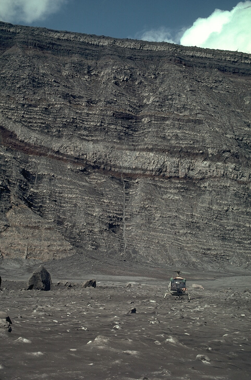 A helicopter on the floor of the inner caldera of Karkar volcano with the 300-m-high caldera wall behind it. The wall was stripped of vegetation by a powerful phreatic explosion on 8 March, revealing the stratigraphy of lighter-colored lava flows and darker pyroclastic deposits that form the edifice. This location is covered by ash and blocks from the March 8 explosion that occurred one month earlier. Photo by William Melson, 1979 (Smithsonian Institution)