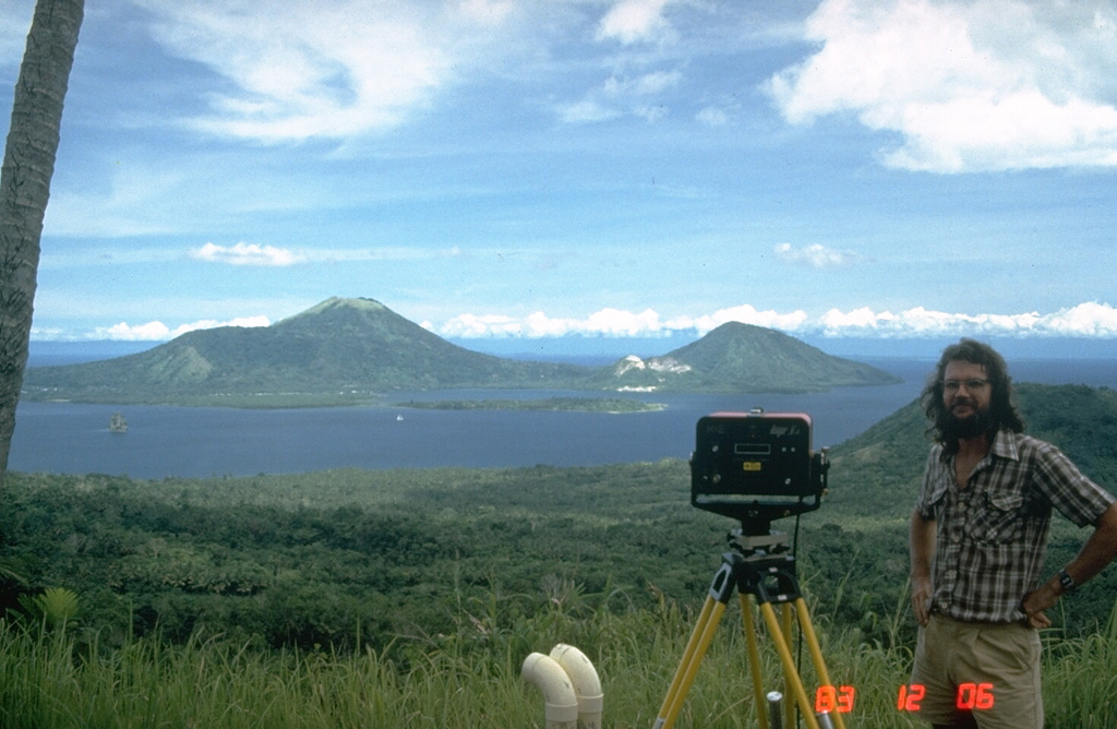 A volcanologist from the Rabaul Volcano Observatory beside an instrument used to make electronic distance measurements (EDM) across Rabaul caldera. Repeated precise measurements of the distance to stations on opposite sides of the caldera are used for monitoring the slow decade-long deformation that preceded a major eruption in 1994. Two pre-caldera peaks, Mount Kombiu (left) and Mount Turanguna (right) are located near the NE caldera rim. Photo by Norm Banks, 1983 (U.S. Geological Survey).