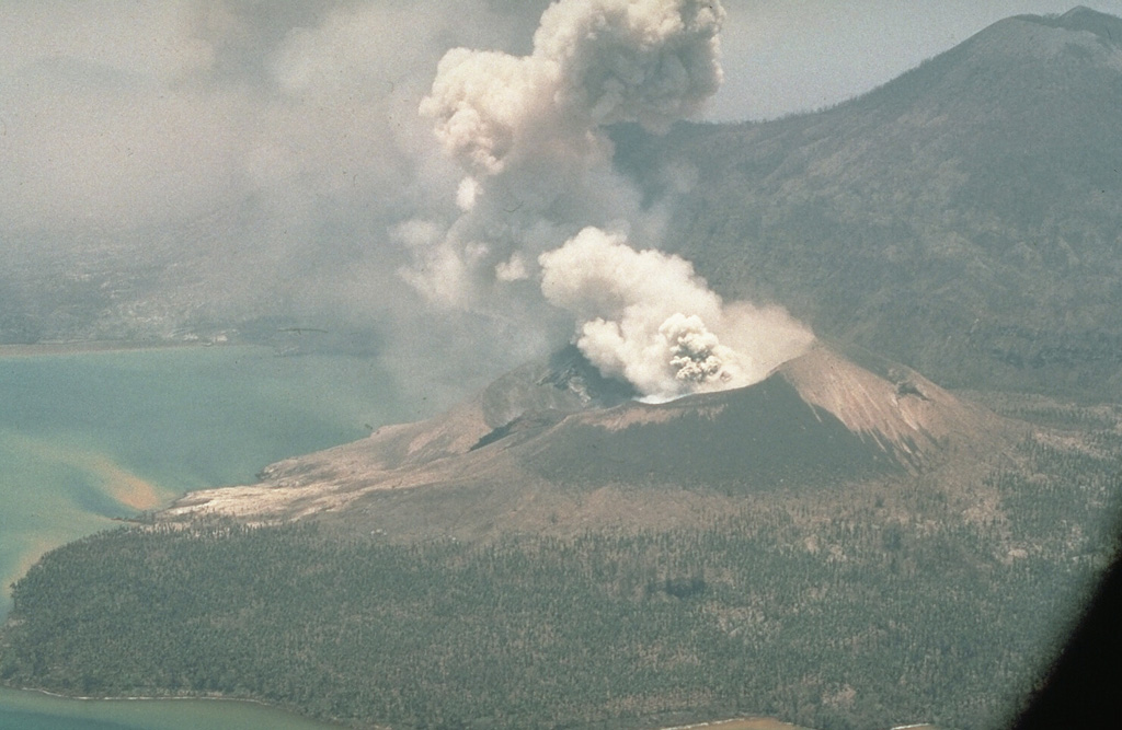 Tavurvur ejects an ash plume in October 1994, following an eruption that began on 19 September. It is seen here from a helicopter with Kombiu volcano (upper right) to the N. Intermittent eruptive activity continued at Tavurvur for several years after the 1994 eruption. Photo by Elliot Endo, 1994 (U.S. Geological Survey).