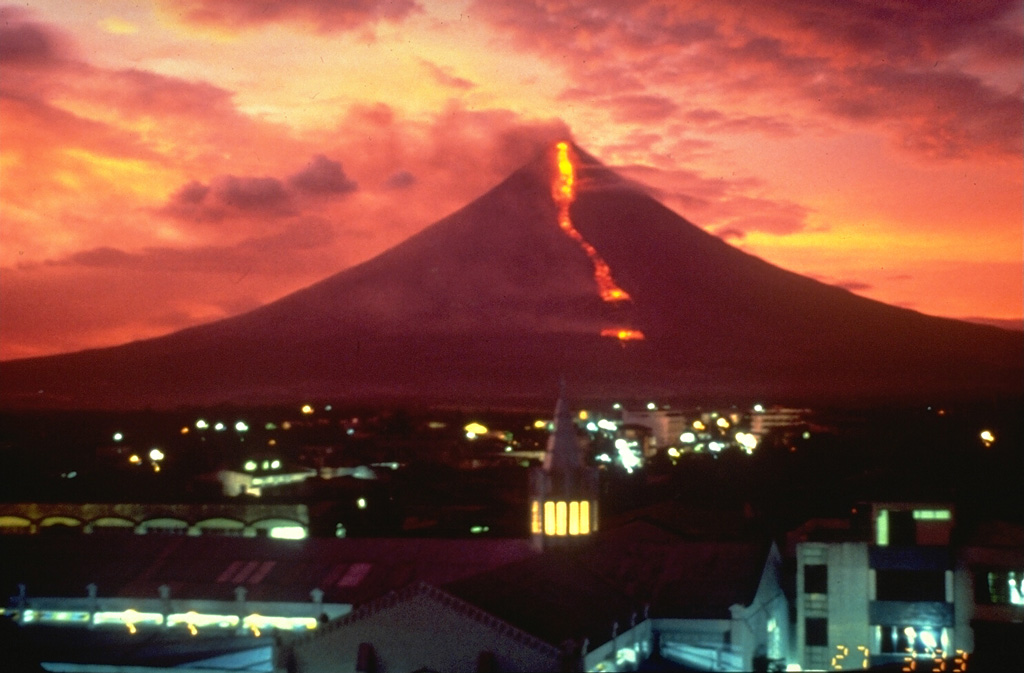 An incandescent lava flow, seen in this sunset view from Legaspi City, descends the SE flank towards the village of Buyuan on 27 March 1993. The eruption began on 2 February with an explosion and pyroclastic flow that traveled 6 km down the E flank and killed 75 people. During the rest of the eruption, which ended in early April, slow lava effusion was accompanied by small pyroclastic flows and ash emissions. Photo by Philippine Institute of Volcanology and Seismology, 1993.