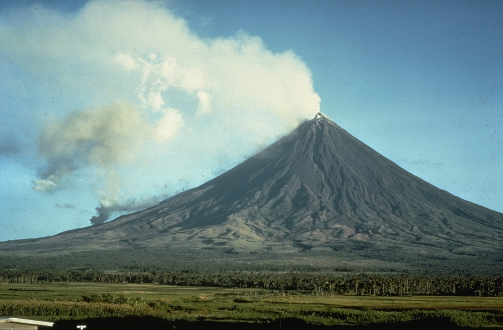 A gas-and-steam plume rises from the summit of Mayon volcano in this May 1968 view from Legaspi City on the SSE flank. The smaller plume on the lower left flank is from the lava flow front that descended 4 km from the summit. Photo by Jim Moore, 1968 (U.S. Geological Survey).