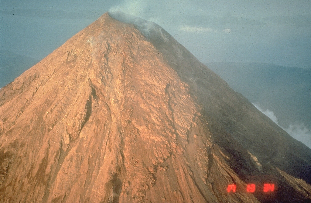 The upper flanks of the symmetrical Mayon are exceptionally steep, averaging 35-40 degrees. They are covered with solidified lava agglutinate, the molten ejecta of lava fountains. This 1984 photo from the WSW shows a 250-m-deep, 3-km-long gully in the shadow at the right that was eroded by pyroclastic flows during the 1984 eruption. Photo by Chris Newhall, 1984 (U.S. Geological Survey).