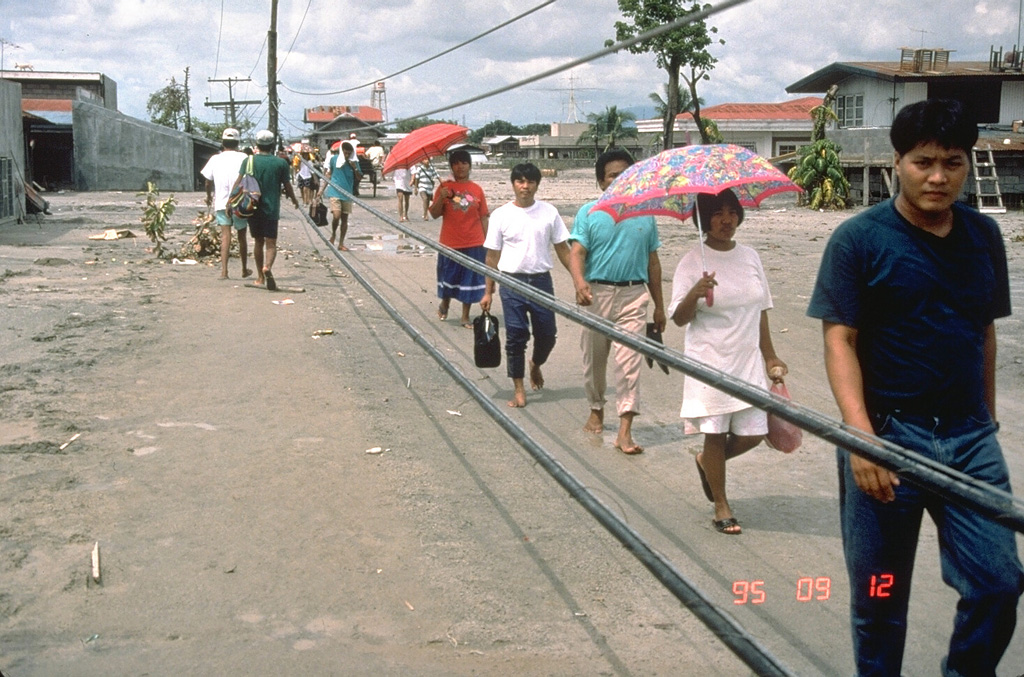 Residents of the city of Bacolor in the Philippines, 38 km SE of Pinatubo, persevere in the face of widespread devastation from lahars. They are walking on the surface of a solidified 5-m-deep deposit of volcanic debris next to wires that are the original electrical power lines formerly high above the street level. Houses and businesses in the background of this September 1995 photo are buried to 2nd-floor levels. Photo by Chris Newhall, 1995 (U.S. Geological Survey).