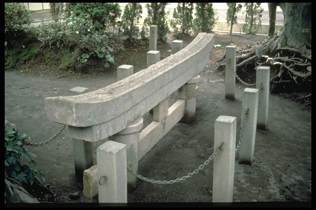 This torii, a Shinto shrine gate, was partially buried by ashfall from the 1914 eruption. The posts and chains surround the gate to protect this historical landmark. Originally 3 m high, the torii is located at the village of Kurokami on the E coast of Sakurajima, 5 km from the summit. Photo by Norm Banks, 1981 (U.S. Geological Survey).