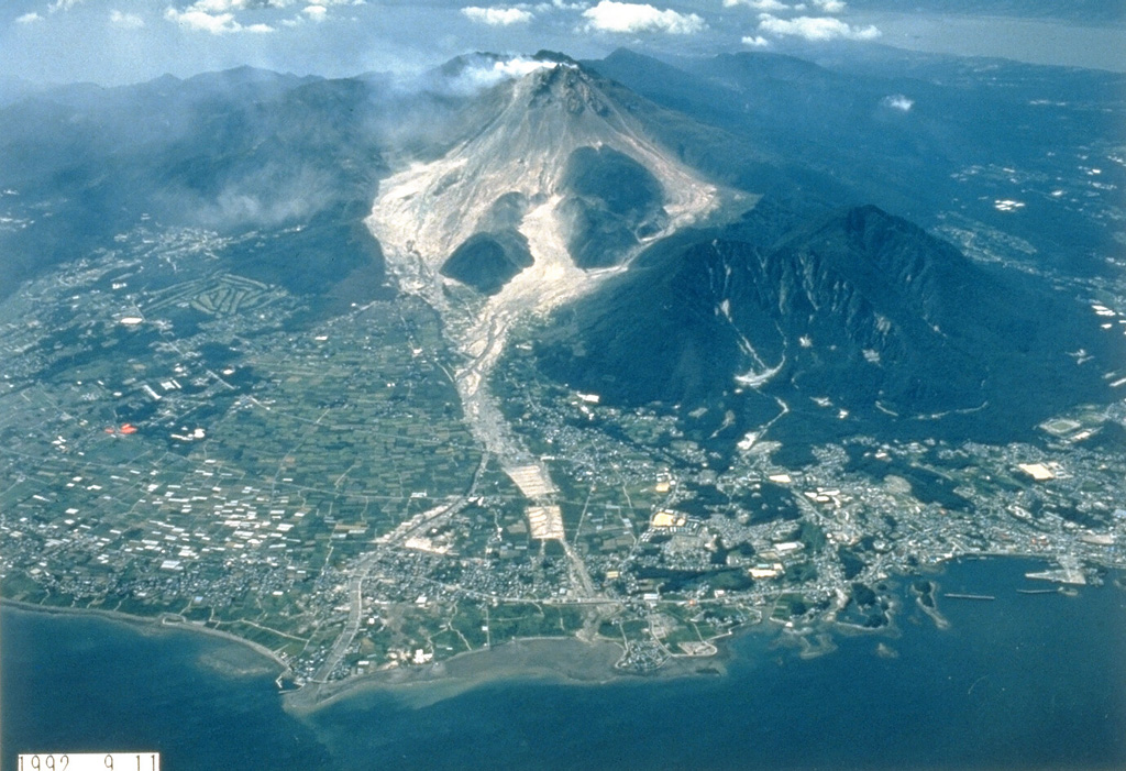 Light-colored block-and-ash flow deposits below the growing Fugendake lava dome are shown in this 11 September 1992 view from the E. Explosive activity began in November 1990 and preceded lava dome growth that began in May 1991. This was accompanied by pyroclastic flows that continued until February 1995 and caused disruption for villages and towns on the outskirts of Shimabara City in the foreground. The two lava domes of Shichmenzan and Mayuyama form the vegetated peaks to the right. Photo courtesy Takashi Yamada, 1992 (Public Works Research Institute, Ministry of Construction).