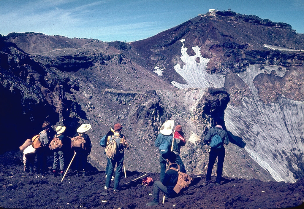 Hikers peer into the 700-m-wide summit crater of Mount Fuji from its E rim. More than 100,000 people ascend its slopes yearly during the 2-month summer climbing season. A white meteorological observatory (upper right) sits at the summit, which is 240 m above the crater floor. A red oxidized scoria layer across the summit crater rim was emplaced about 2,100 years ago. Photo by Lee Siebert, 1963 (Smithsonian Institution).