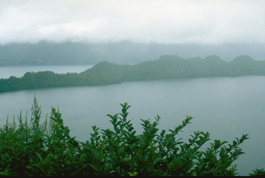 The peninsula across the center of the photo is the SW rim of Nakanoumi caldera. The 3-km-wide caldera formed about 5,400 years ago during the largest Towada Holocene eruption . The eruption was followed by the collapse of the Goshikiiwa cone at the southern end of the caldera. Clouds obscure the western caldera rim in the background. Photo by Lee Siebert, 1977 (Smithsonian Institution).
