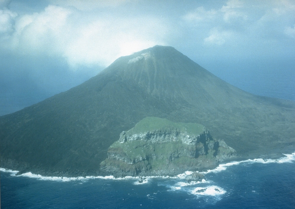 The 2-km-wide island of Farallon de Pajaros is the northernmost and most active volcano of the Mariana Islands. It has been referred to as the "Lighthouse of the western Pacific." The symmetrical, sparsely vegetated cone formed within a caldera, remnants of which form the peak seen here in 1980 in the center along the SE coast. Both summit and flank vents have been active during historical time; flank fissures formed lava flows along the coast. Photo by Winfrey, 1980 (U.S. Navy).
