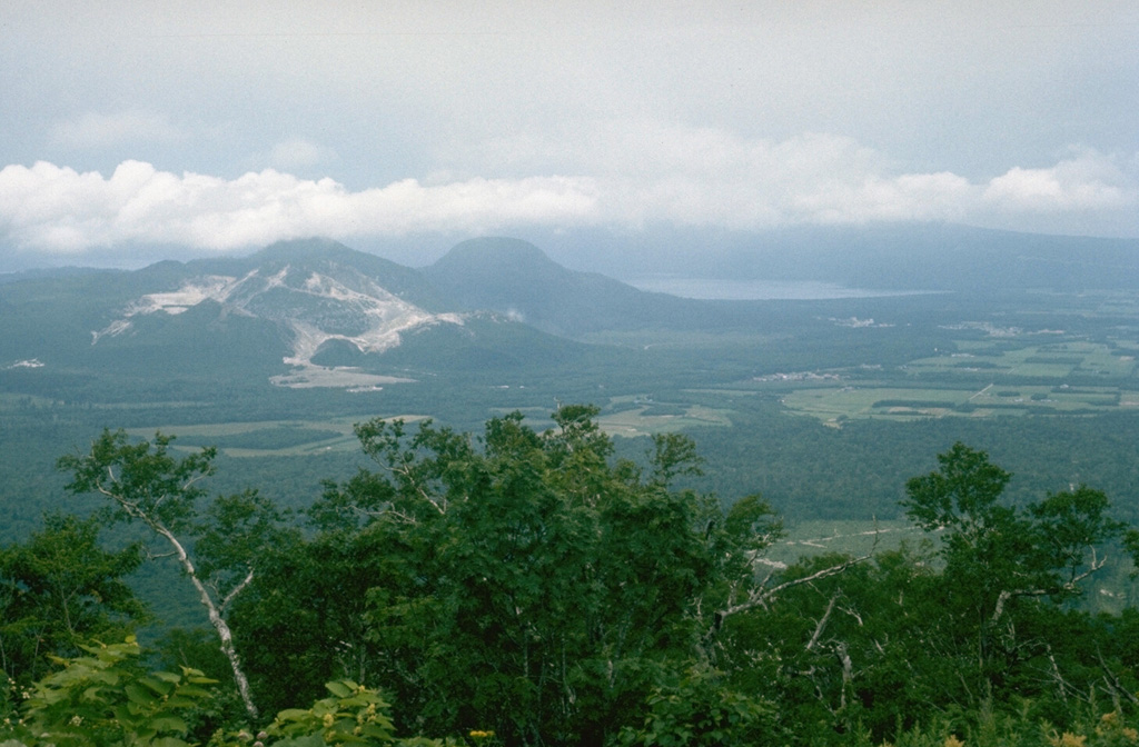 A cluster of lava domes is located in the center of Kussharo caldera, which is partially filled by Lake Kutcharo (upper right). Light-colored areas of hydrothermal alteration cover the slopes of Atosanupuri (left), seen here from the east with the rounded Sawanchisappu lava dome to its right. The far western wall of the 20 x 26 km caldera lies in the clouds at the far end of the lake. Photo by Lee Siebert, 1977 (Smithsonian Institution).
