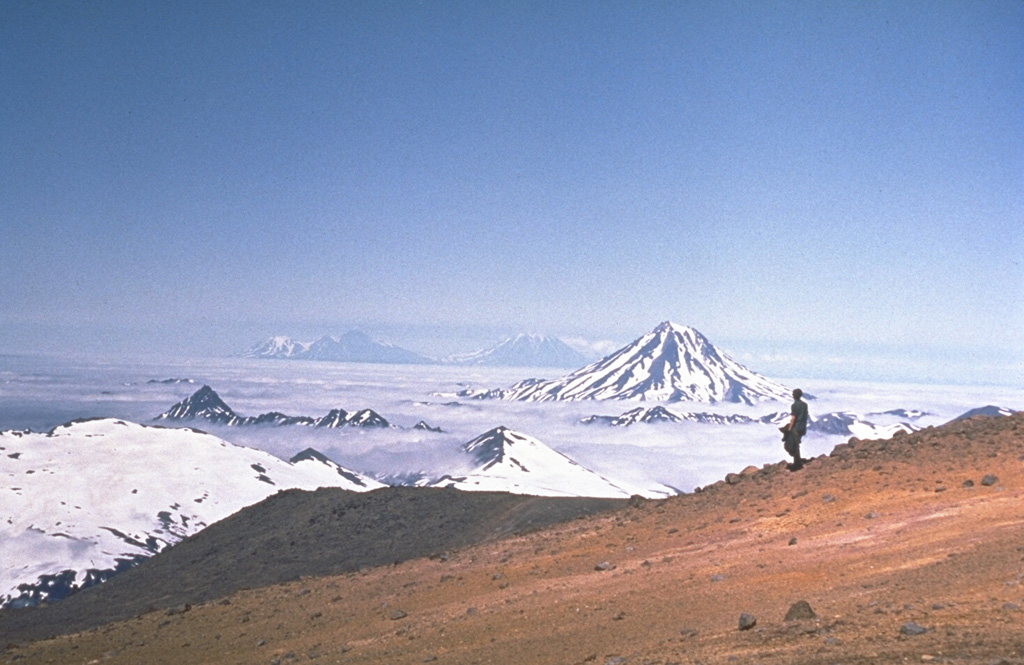 Vilyuchik volcano to the right is located NE of Mutnovsky volcano. Clouds fill one of the snow-mantled summit craters of the Mutnovsky, one of the most active volcanoes of southern Kamchatka. Koryaksky (left) and Avachinsky (right) volcanoes rise in the far-distance across cloud-covered Avachinsky bay. Photo by Oleg Volynets, 1971 (Institute of Volcanology, Petropavlovsk).