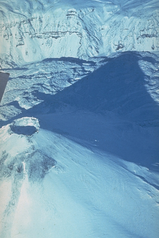 The shadow of Karymsky is seen here across lava flows within the caldera in which it was constructed. The NW caldera wall forms the 300-m-high cliff in the background. The 5-km-wide caldera formed during one of Kamchatka's largest Holocene eruptions about 7,500 radiocarbon years ago. Karymsky was constructed during frequent explosive and effusive eruptions during the past 6,000 years. Its recent eruptive period began about 500 years ago. Photo by Yuri Doubik (Institute of Volcanology, Petropavlovsk).