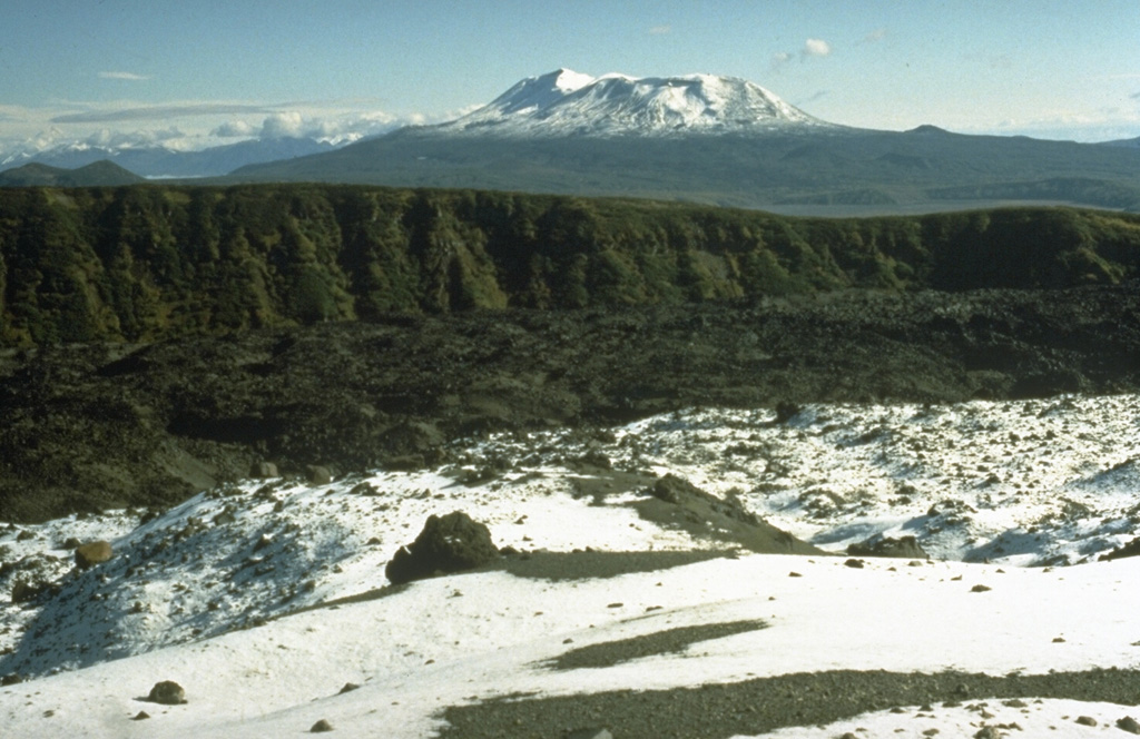 The eastern margin of Karymsky caldera, which was created about 7,500 years ago, forms the steep wall across the center of the photo. Fresh, dark-colored lava flows from Karymsky cover the caldera floor. The snow-covered volcano to the NE is Maly Semyachik, which has also produced frequent historical eruptions. Photo by Dan Miller (U.S. Geological Survey).