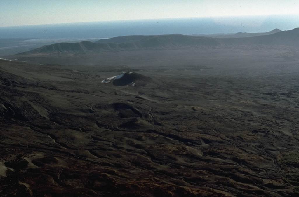 Volcanism along the elongated Maly Semiachik massif has progressively migrated to the south. The southern flank is mantled by Holocene cones and lava flows. The SE rim of a large Pleistocene caldera forms the low ridge in the background and the Pacific Ocean is visible in the distance. Photo by Dan Miller, 1990 (U.S. Geological Survey).