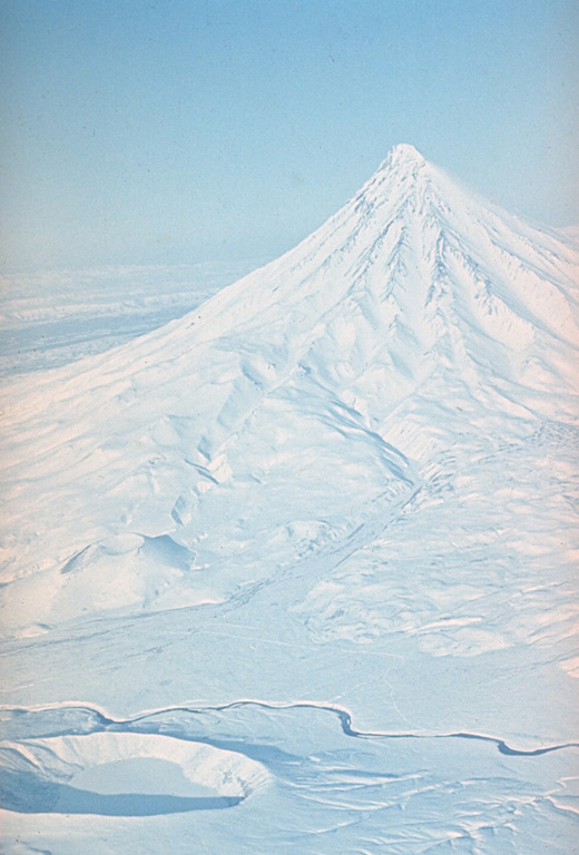 Kronotsky rises NE of the Kronotsky River, shown across the bottom of the photo. A small cone above the river to the right is one of several on the SW flank. The crater to the lower right is a lake-filled maar that formed at the northern end of a rift zone extending 8 km NNE from Krasheninnikov. The maar erupted along the NE flank of a large Pleistocene caldera inside which Krasheninnikov formed. Photo by Yuri Doubik (Institute of Volcanology, Petropavlovsk).