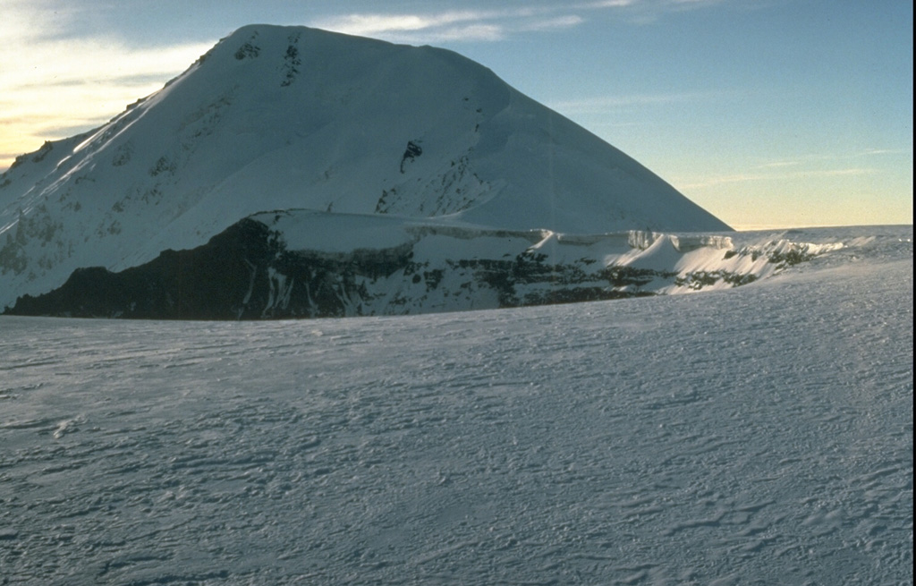 The summit of Plosky Tolbachik contains a 3-km-wide caldera whose floor is covered by the glacier in the foreground. It contains a nested inner 1.8-km-wide caldera (center) with a crater that increased substantially in size during the 1975-76 eruption. Ostry Tolbachik stratovolcano towers west of the inner caldera of Plosky Tolbachik in this 1990 view. Photo by Dan Miller, 1990 (U.S. Geological Survey).