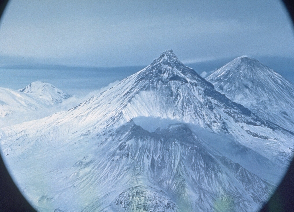 Four volcanoes of the Klyuchevskaya volcano group are visible in this north-looking view. A gas plume emanates from the summit of Bezymianny in the foreground, which is dwarfed by Kamen behind it. Klyuchevskoy is to the upper right, and Ushkovsky is on the left horizon. Krestovsky forms the rounded summit and the glacier-covered Ushkovsky caldera is visible to the far-left. Photo by Oleg Volynets (Institute of Volcanology, Petropavlovsk).