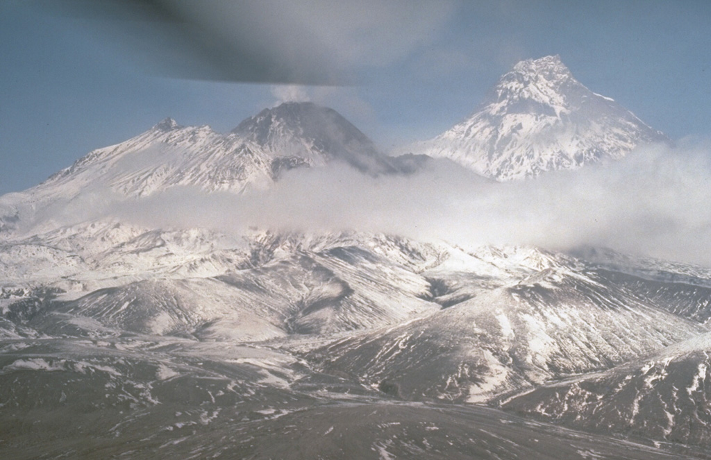 A weak plume rises from the summit lava dome of Bezymianny in September 1990. This view from the south shows Kamen to the right; to the left are the snow-mantled slopes of the outer flanks of the pre-1956 Bezymianny edifice. Collapse of the summit during a catastrophic eruption that year produced a large horseshoe-shaped crater that has subsequently been largely filled by growth of the lava dome. Photo by Dan Miller, 1990 (U.S. Geological Survey).