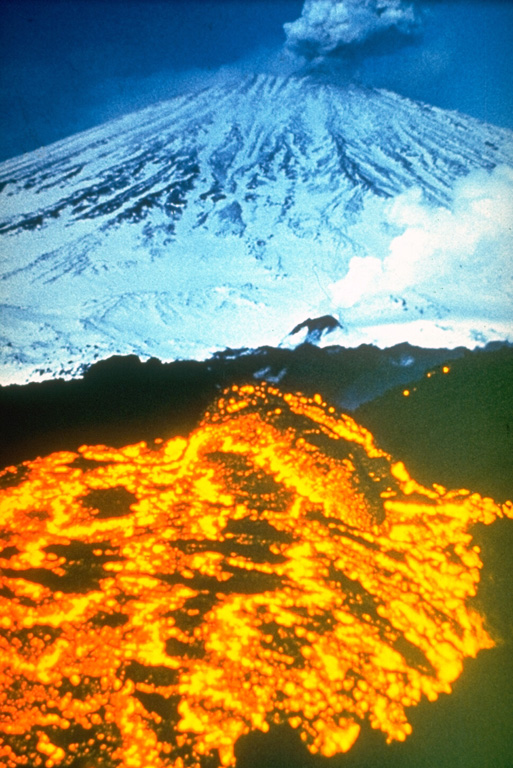 An incandescent lava flow erupts from an E-flank vent on Klyuchevskoy in 1983 while an ash plume rises from the summit crater. An eruption from an east flank fissure began on 8 March 1983. A scoria cone that formed during the initial stage was the source of the flank activity. Strombolian eruptions during the main phase were followed by lava emission at varying rates. Flank activity was preceded by summit crater gas-and-ash emissions beginning 7 October 1982. Photo by Yuri Doubik, 1983 (Institute of Volcanology, Petropavlovsk).