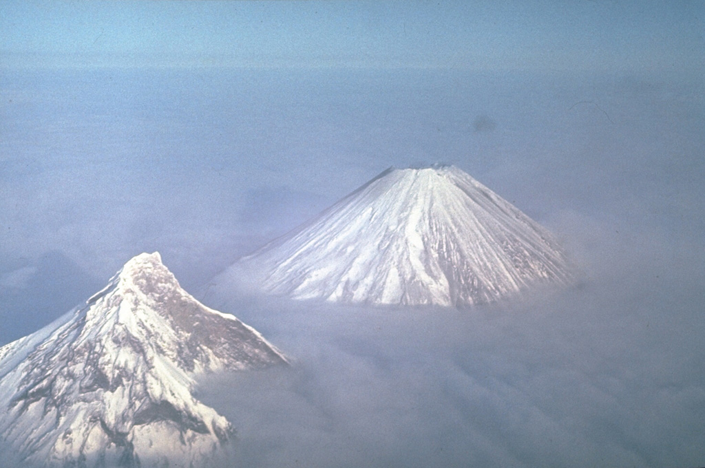 Two of Kamchatka's highest volcanoes rise above the clouds. Their differing morphologies reflect contrasting geologic histories. Construction of extensively eroded Kamen volcano (left) took place during the Pleistocene. It has been relatively inactive since. Its eastern (right) side was removed by a massive landslide about 1,200 years ago, leaving the steep escarpment. Symmetrical Klyuchevskoy, in contrast, is one of Kamchatka's youngest and most active volcanoes. Photo by Yuri Doubik (Institute of Volcanology, Petropavlovsk).