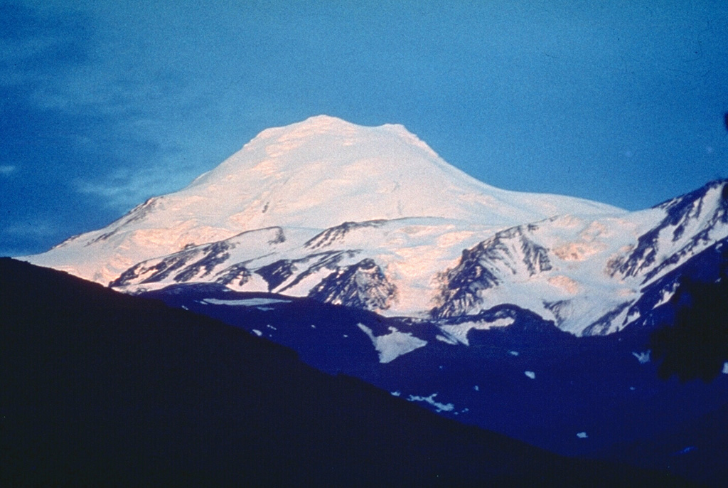 The setting sun gilds the SW flank of Ichinsky, the most prominent volcano of the Sredinny Range. Two glacier-covered lava domes form the summit. The massive stratovolcano is one of the largest volume volcanoes in Kamchatka. No historical eruptions are known from Ichinsky, but Holocene eruptions have produced dactic-to-rhyodacitic lava domes and voluminous basaltic-to-andesitic lava flows from flank vents. Photo by Oleg Volynets, 1977 (Institute of Volcanology, Petropavlovsk).