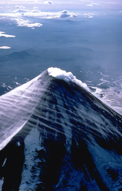 The small summit crater of symmetrical Shishaldin volcano produces an almost constant steam plume.  Strong winds in this 1978 view from the east also sweep streams of linear atmospheric clouds across the flanks of the volcano.  The dominantly basaltic volcano is built atop an older volcano, remnants of which can be seen on the west and NE sides. Copyrighted photo by Katia and Maurice Krafft, 1978.