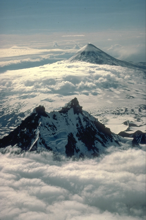 The effects of differential erosion are dramatically seen in this aerial view of two Alaskan volcanoes.  Isanotski volcano in the foreground, sometimes referred to as "Ragged Jack," has been extensively dissected by glaciers that have excavated steep walls separated by narrow ridges.  The symmetrical Shishaldin volcano in the background is sometimes referred to as the Mount Fuji of the Aleutians.  In contrast to Isanotski, Shishaldin is one of Alaska's most active volcanoes.  Frequent eruptions maintain the young, constructional profile of the volcano. Copyrighted photo by Katia and Maurice Krafft, 1978.