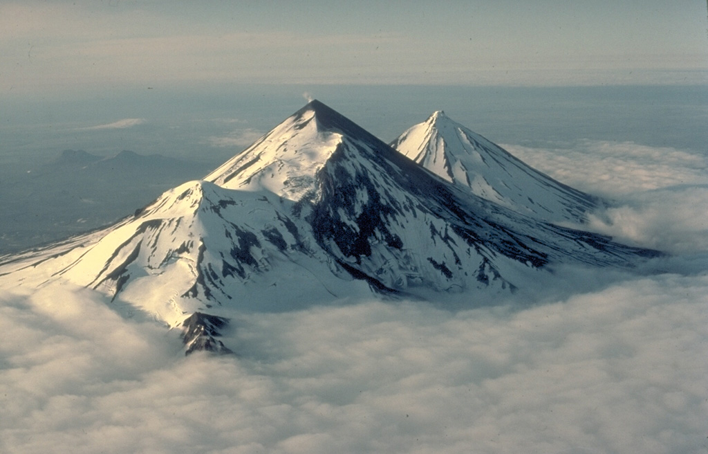 The twin volcanoes of Pavlof (left) and Pavlof Sister (right) rise above a sea of clouds on the southern Alaska Peninsula.  The two conical stratovolcanoes are constructed on the same NE-SW line as Little Pavlof, a flank cone in the left center of this 1978 view from the SW.  Pavlof, its NE slopes darkened by ash, is the most active volcano of the Aleutian arc.  Ash-free Pavlof Sister is somewhat more glacially dissected than Pavlof and has had only one historical eruption. Copyrighted photo by Katia and Maurice Krafft, 1978.