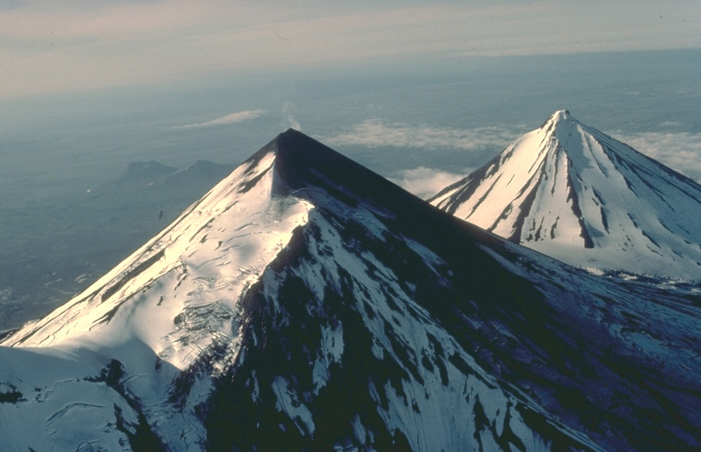 The twin volcanoes of Pavlof (left) and Pavlof Sister (right) are prominent landmarks on the southern Alaska Peninsula.  The summits of the two conical stratovolcanoes are only 5 km apart.  This 1978 view from the SW shows a steam plume rising from the 2519-m summit of Pavlof, the most active volcano of the Aleutian arc.  Pavlof Sister, somewhat more eroded than Pavlof, has had only a single historical eruption, in the 18th century. Copyrighted photo by Katia and Maurice Krafft, 1978.