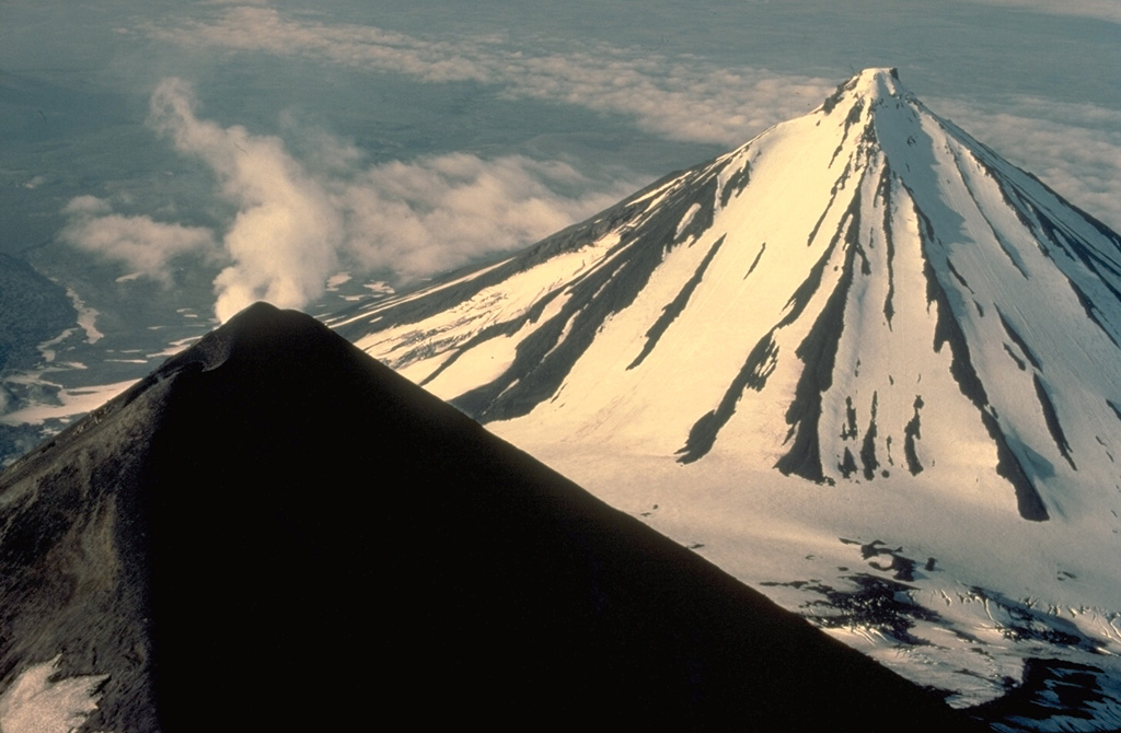 A steam plume rises from the summit of Pavlof volcano, whose upper slopes are snow-free as a result of its frequent eruptions.  More-extensive glacial dissection of Pavlof Sister volcano (upper right) has produced a series of parasol-ribbed ridges that radiate from the summit.  Only one historical eruption is known from Pavlof Sister. Copyrighted photo by Katia and Maurice Krafft, 1978.