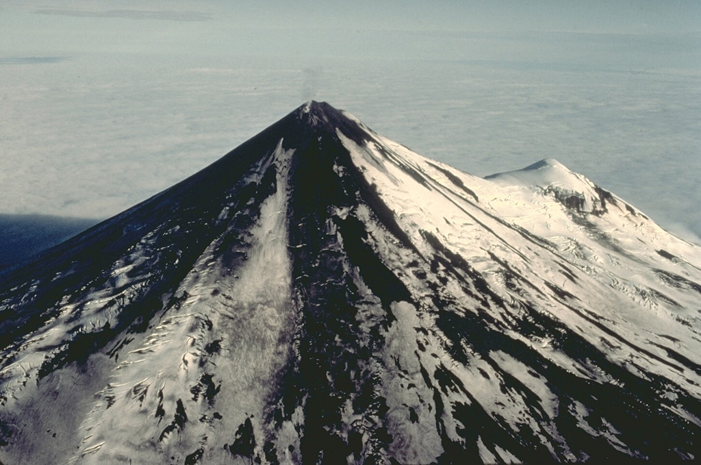 Pavlof volcano is the most active of the Aleutian arc and one of the most prominent landmarks of the southern Alaska Peninsula.  A small steam plume rises from the 2519-m summit of Pavlof in this aerial view from the NW with the flank cone of Little Pavlof on the right.  The conical volcano was constructed NE of the rim of Emmons Lake caldera and has active vents high on the north and east sides near the summit.  Its frequent eruptions are typically strombolian and vulcanian in character, with occasional spatter-fed lava flows. Copyrighted photo by Katia and Maurice Krafft, 1978.