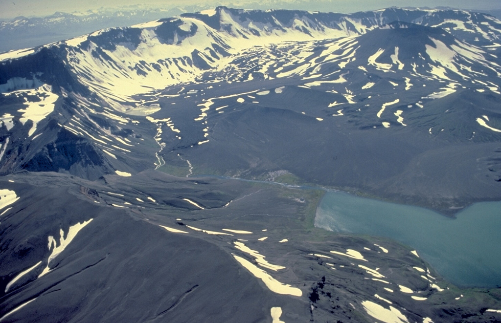The 10-km-wide Aniakchak caldera is one of the most dramatic volcanoes on the Alaska Peninsula.  It was formed about 3400 years ago during an eruption in which voluminous pyroclastic flows reached the Bering Sea, 80 km away.  This view from the NE shows the largest post-caldera volcano, Vent Mountain (upper right) and Surprise Lake (lower right), which drains through The Gates (left center), a steep-walled notch in the 1-km-high eastern caldera rim.  The only historical eruption of Aniakchak took place in 1931. Copyrighted photo by Katia and Maurice Krafft, 1978.