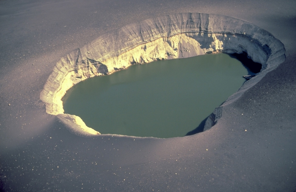 The 300-m-wide East Maar is one of the two Ukinrek Maars, which were formed during an eruption in 1977 through glacial deposits on the Alaska Peninsula.  This fresh maar, in contrast to older ones whose rims have been modified by slumping and erosion, has steep-sided walls.  The explosions blasted through the pre-existing, light-colored glacial till forming the bottom half of the distant crater wall and then deposited the dark, bedded layers in the upper half of the crater wall as well as the dark blanket of blocks and ash surrounding the maar. Copyrighted photo by Katia and Maurice Krafft, 1978.