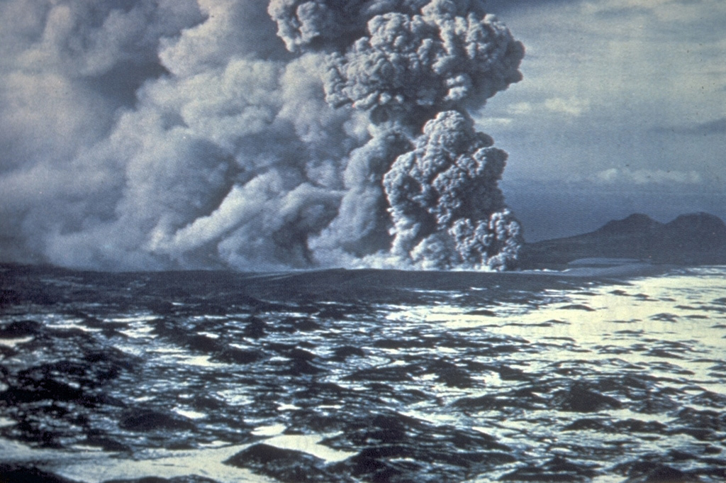 Phreatomagmatic eruptions produced when magma came in contact with groundwater eject an ash plume at the Ukinrek Maars on the Alaska Peninsula. This photo from the south on 5 April 1977 shows explosive eruptions from East Maar, one of two new craters that formed in glacial sediments, in an area without previous volcanic activity. The hummocky, snow-covered surface in the foreground is a debris avalanche deposit from Peulik. Photo by Jim Faro (Alaska Department of Fish and Game).