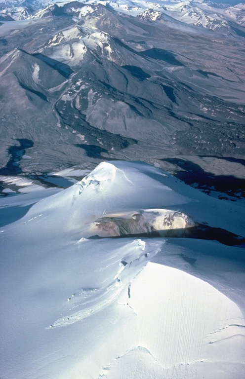 The summit crater of glacier-covered Mount Mageik appears in the foreground of this 1978 view looking NE across Katmai Pass to Trident volcano.  These two volcanoes stand guard at the head of the Valley of Ten Thousand Smokes.  Mageik's young crater contains a shallow turbulent lake.  The fresh, black lava flows descending the SW flank of Trident volcano were emplaced in a series of eruptions during 1953-1966. Copyrighted photo by Katia and Maurice Krafft, 1978.