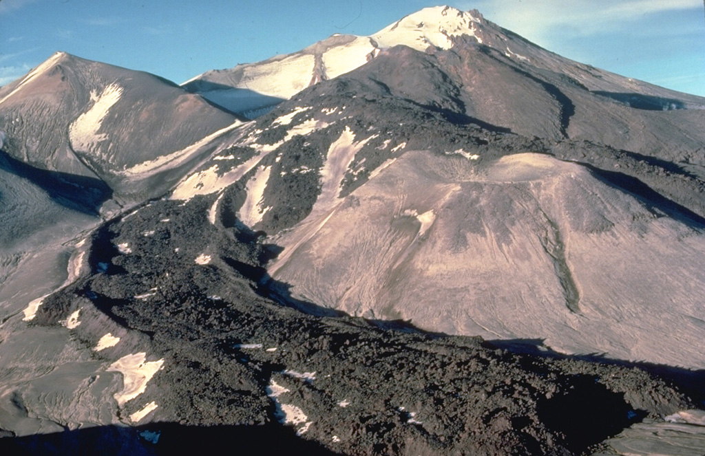 A blocky andesitic lava flow, its margins marked by prominent flow levees, descends the SW flank of Trident volcano in Katmai National Park, Alaska.  The lava flow, seen here from the WSW in 1978, was part of a series of eruptions from 1953 to 1968 that constructed the snow-covered cone in the background.  The lava flows diverge around the lighter-colored surface of an older lava dome at the right center. Copyrighted photo by Katia and Maurice Krafft, 1978.