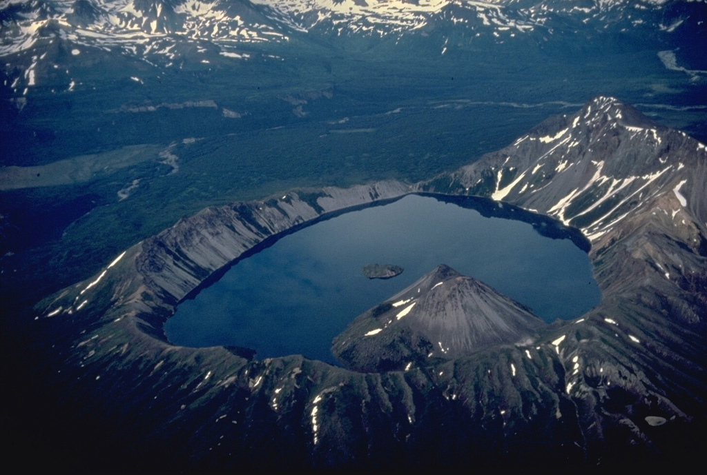 Kaguyak caldera was formed following powerful dacitic explosive eruptions about 5800 years ago.  An extensive apron of pyroclastic-flow deposits surrounds the 2.5-km-wide caldera.  This view from the SW shows two post-caldera lava domes that form a peninsula into the lake and another lava dome that forms a small island that rises about 10 m above the center of the lake.  The low notch at the far end of the caldera is a pre-caldera stream valley that was beheaded by caldera formation. Copyrighted photo by Katia and Maurice Krafft, 1978.