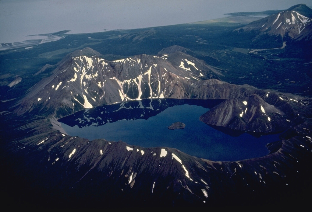 The small, but dramatic Kaguyak caldera on the northern Alaska Peninsula contains a lake more than 180-m deep whose surface is 550-m below the caldera rim.  Two post-caldera lava domes form the large peak that rises above the rim of the 2.5-km-wide caldera at the right.  The small island in the center of the lake is another lava dome.  Collapse of the caldera occurred following powerful dacitic explosive eruptions about 5800 years ago.  Shelikof Strait is visible at the top of the photo. Copyrighted photo by Katia and Maurice Krafft, 1978.