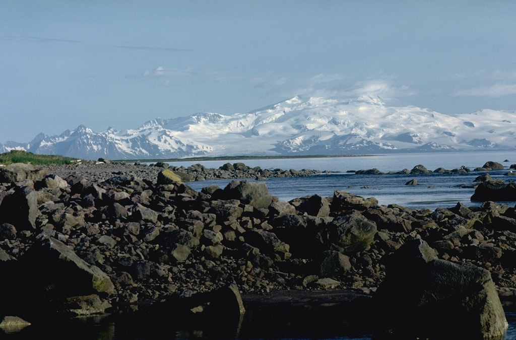 The broad, glacier-covered Mount Douglas rises across Kamishak Bay as seen from Augustine Island to its NE. The rocks in the foreground are part of a young debris avalanche deposit from Augustine. Photo by Lee Siebert, 1986 (Smithsonian Institution).
