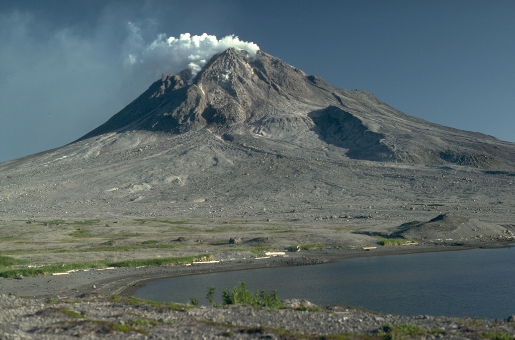 Several features from the Augustine 1883 eruption can be seen in this 1987 photo of the N flank. The hill across the shallow lagoon to the lower right is a debris avalanche hummock produced by collapse of the summit and N flank in 1883. The result of the avalanche was a large open crater that is largely filled by 20th-century lava domes; the outer wall is in the shadow to the left. The area covered with sparse green vegetation is a pyroclastic flow deposit from the 1883 eruption. Photo by Lee Siebert, 1987 (Smithsonian Institution).