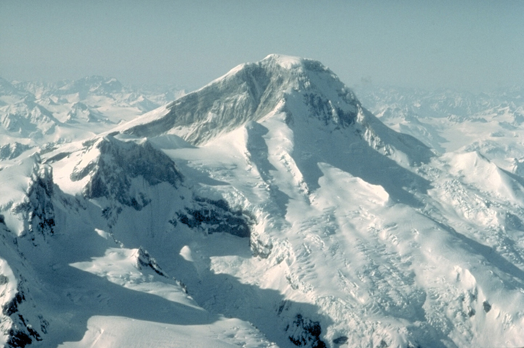 Iliamna volcano, seen here from the SE, is an ice-clad stratovolcano that has been extensively eroded by glaciers.  Four peaks occur along a 5-km-long, N-S-trending summit ridge, ending with North Twin and South Twin peaks at the left center.  No major Holocene tephras have been identified from Iliamna.  Many reports of historical eruptions have mistaken powerful episodes of steam emission from fumaroles high on the south and east flanks. Copyrighted photo by Katia and Maurice Krafft, 1986.