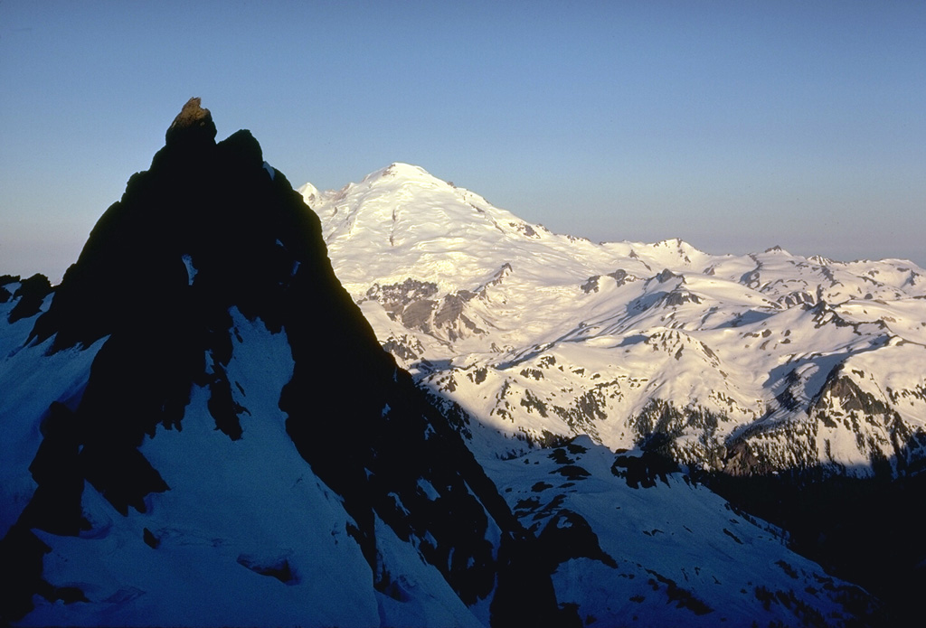 The glaciated surface of Mount Baker is seen here from neighboring Mount Shuksan. The peak in the shadow consists of metamorphosed basaltic rocks of the Shuksan Greenschist. The area to the right is the recently discovered Pleistocene Kulshan Caldera, which preceded the construction of Mount Baker. The 4.5 x 8 km caldera is largely filled by up to 1 km of non-welded tuffs and is capped by lava flows.  Photo by Lee Siebert, 1971 (Smithsonian Institution).