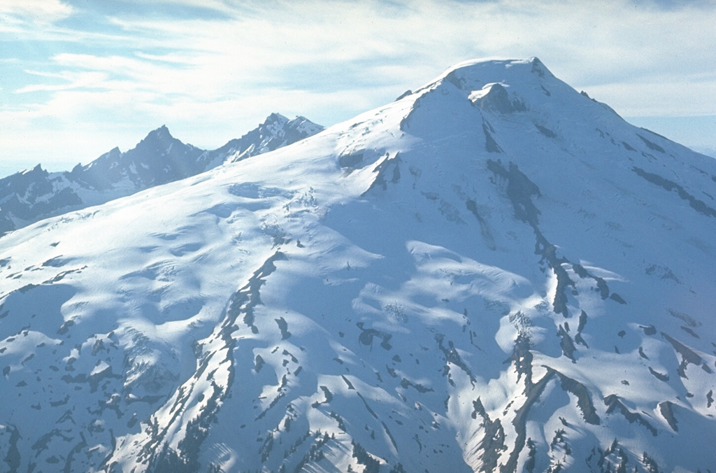 This view from the SE shows the extensive glacial cover on Mount Baker in 1972. Twelve glaciers cover the volcano with 114 km2 of ice, which is the largest glacial icecap of any Cascade volcano, including Mount Rainier. Part of Sherman Crater, the historically active vent, is lit by the sun just below the summit, to the right of Sherman Peak. The eroded Black Buttes volcano is in the background. Photo by Lee Siebert, 1972 (Smithsonian Institution).