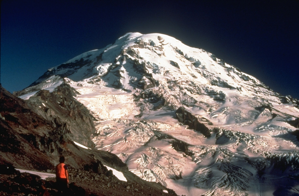 A volcanologist from the U.S. Geological Survey observes the glaciated NW flank of Mount Rainier during a field survey to conduct monitoring measurements on Ptarmigan Ridge. The North Mowich Glacier in the center of the photo descended to about 1,500 m elevation when this photo was taken in 1983. Photo by Lyn Topinka, 1983 (U.S. Geological Survey).