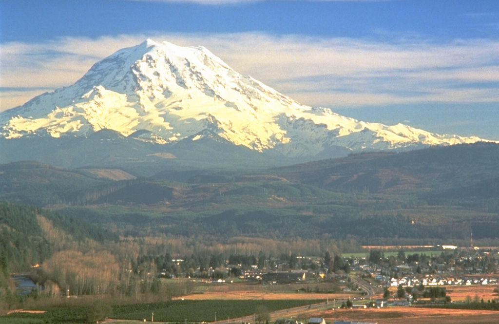 Mount Rainier towers above the town of Orting located 40 km NW in this 1995 photo. The plain underlying the town is composed of the Electron Mudflow, which formed the flat valley floor about 500 years ago. The mudflow, which originated from partial collapse of part of the western flank of Rainier, was about 30 m deep when it exited valleys at the mountain front and flowed onto the Puget Lowland. Photo by Dave Wieprecht, 1995 (U.S. Geological Survey).