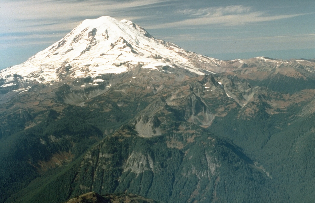 An aerial view from near the eastern margin of Mount Rainier National Park shows the volcano rising above glacially eroded terrain of the Ohanapecosh formation, composed of Tertiary volcanic rocks. The smooth, glaciated upper NE flank in this 1992 photo is the location of the post-collapse cone constructed within the failure scarp left by the Osceola debris avalanche and lahar about 5,600 years ago. Photo by Dave Wieprecht, 1992 (U.S. Geological Survey).