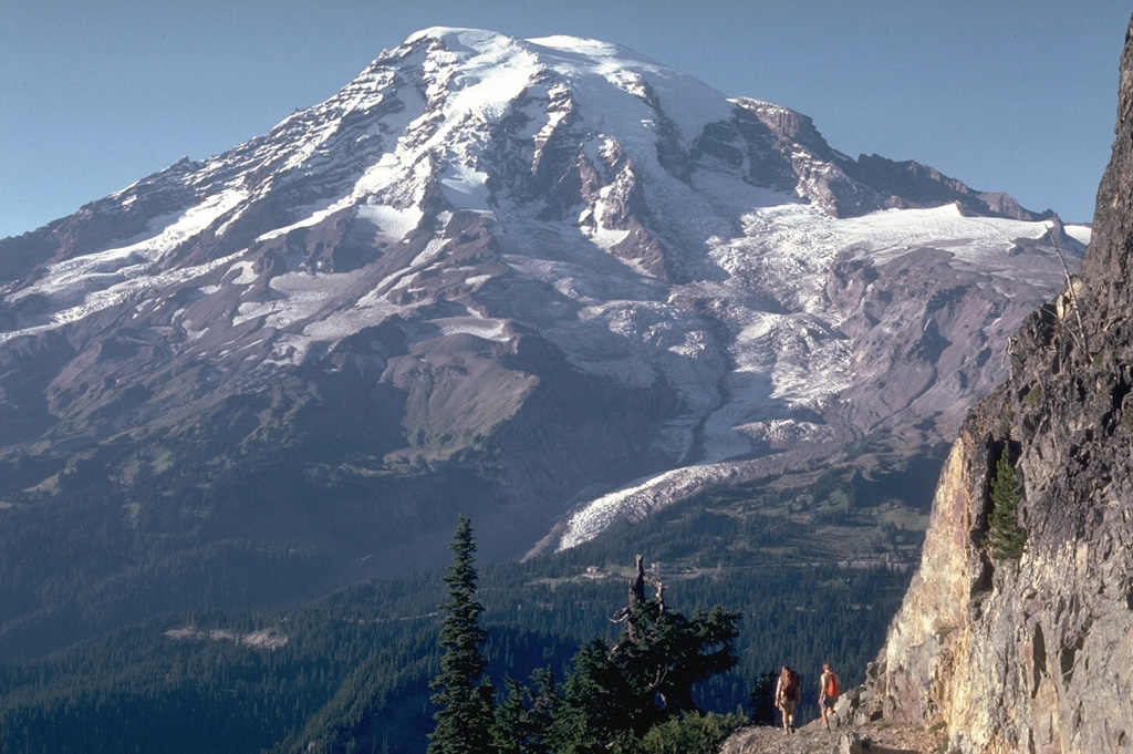 The south flank of Rainier is seen here from the Tatoosh Range, with the Nisqually Glacier below the snow line in this 1980 photo. Meadows and forests of the Paradise area lie immediately below and to the right of the glacier. This is one of 25 named glaciers on Rainier, with the snow, ice, loose rock, and hydrothermal alteration posing a risk of lahars and debris avalanches for surrounding areas. Photo by Lee Siebert, 1980 (Smithsonian Institution).