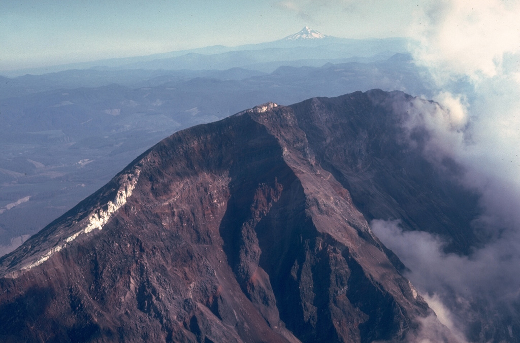 The steep headwall of the scarp created by collapse of St. Helens on 18 May 1980 towers 550 m above the crater floor. The white areas on the crater rim are glaciers that were truncated by the collapse. Steam rises at the right from the new crater in this August 1980 view. Mount Hood is visible in the distance to the south across the Columbia River.  Photo by Lee Siebert, 1980 (Smithsonian Institution).